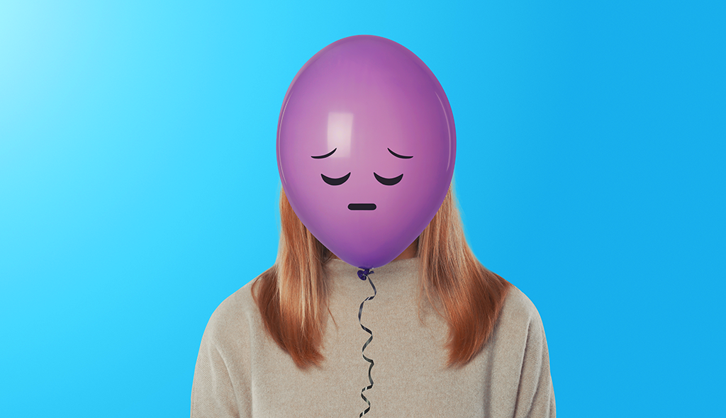 young woman with blonde hair holding a purple sad-faced balloon in front of her face, on a bright blue background.