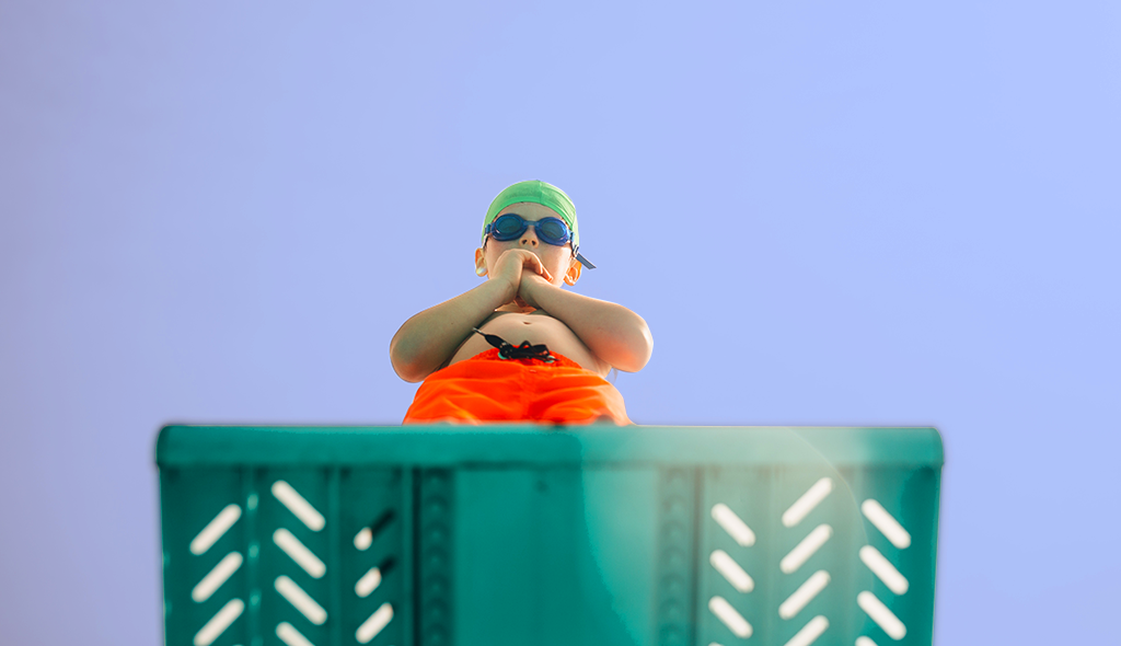 A young boy stands at the edge of a diving board, hesitating to jump, on a light blue-ish purple background.