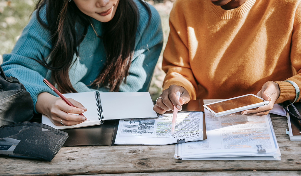 2 young people studying together, writing in notebooks and looking at tablet