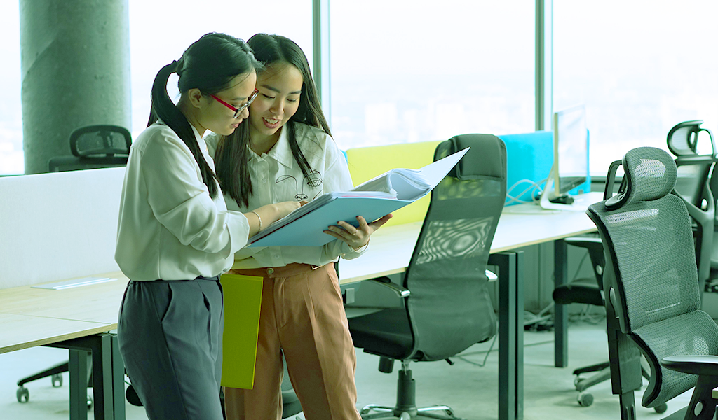 Two young women in a large, bright office space, standing together and looking over a large booklet
