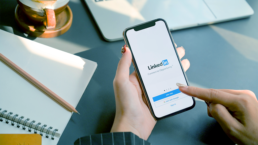 Hands holding a phone, with LinkedIn login screen. Desk, notebook. calendar and coffee in the background.