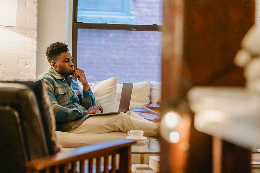 A young man/freelance writer sitting on a couch in his apartment, deep in thought and typing on a laptop computer