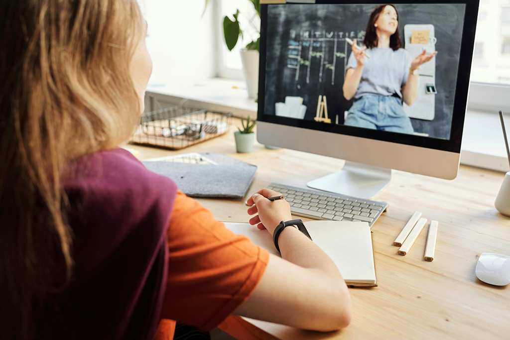 The view over a young woman's shoulder who is sitting at her desk and taking notes while watching an online course